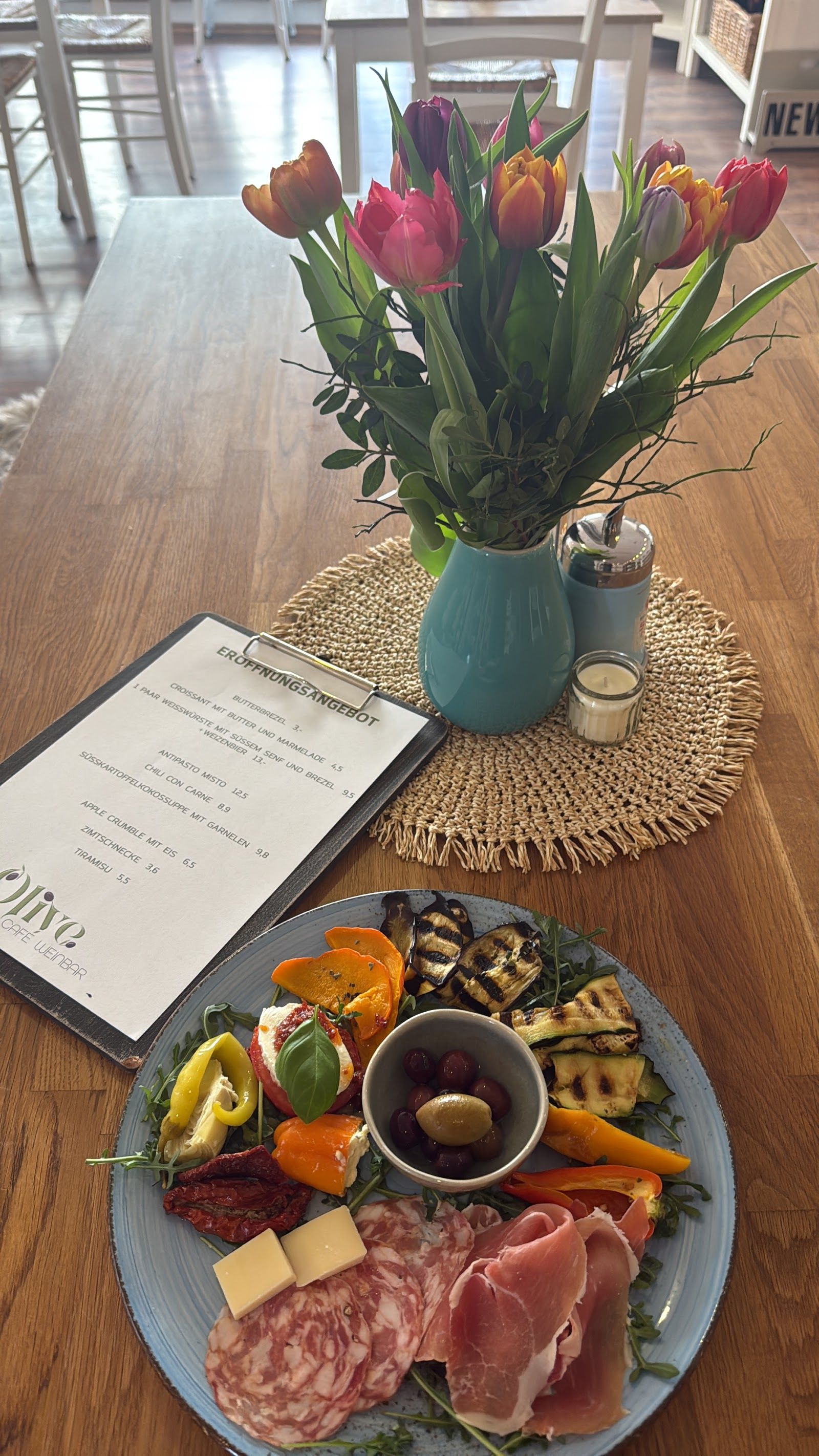 Antipasti-Brett mit der Olive-Menükarte und einem Strauß Tulpen auf hellem Holztisch.