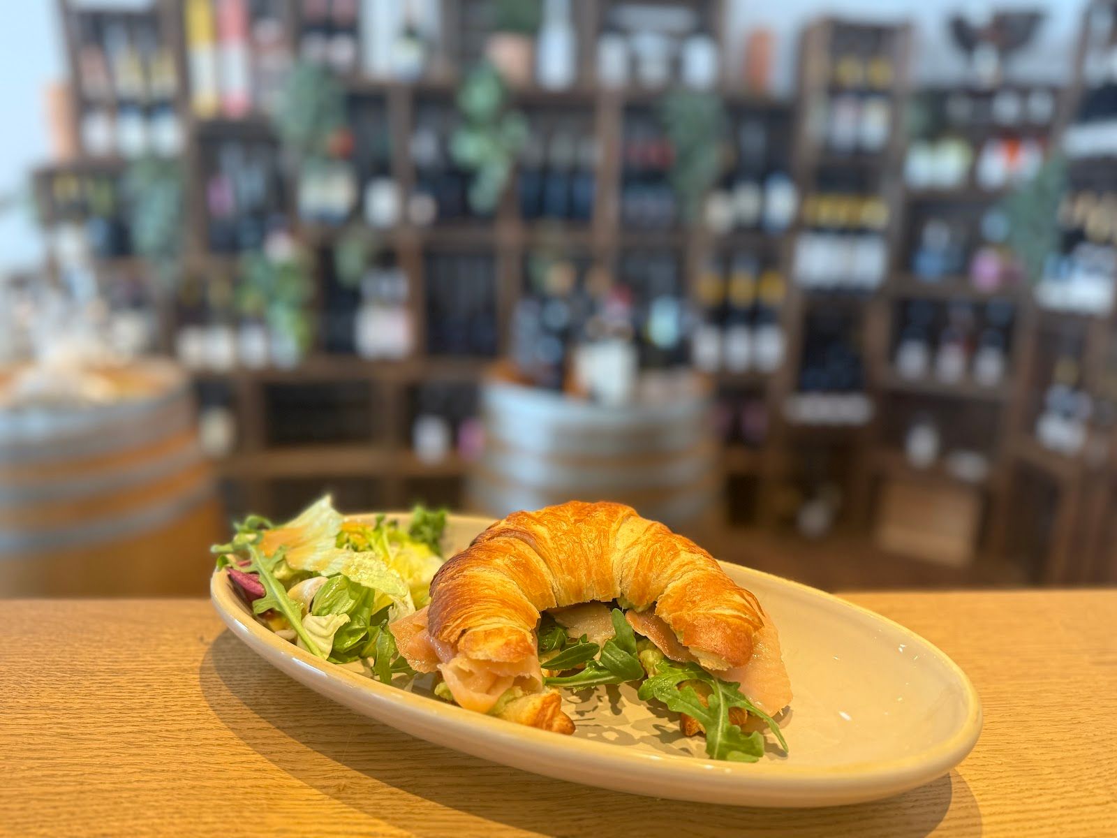 Mit Räucherlachs und Rucola gefülltes Croissant auf cremefarbenem Teller, dahinter die Weinwand von Olive.