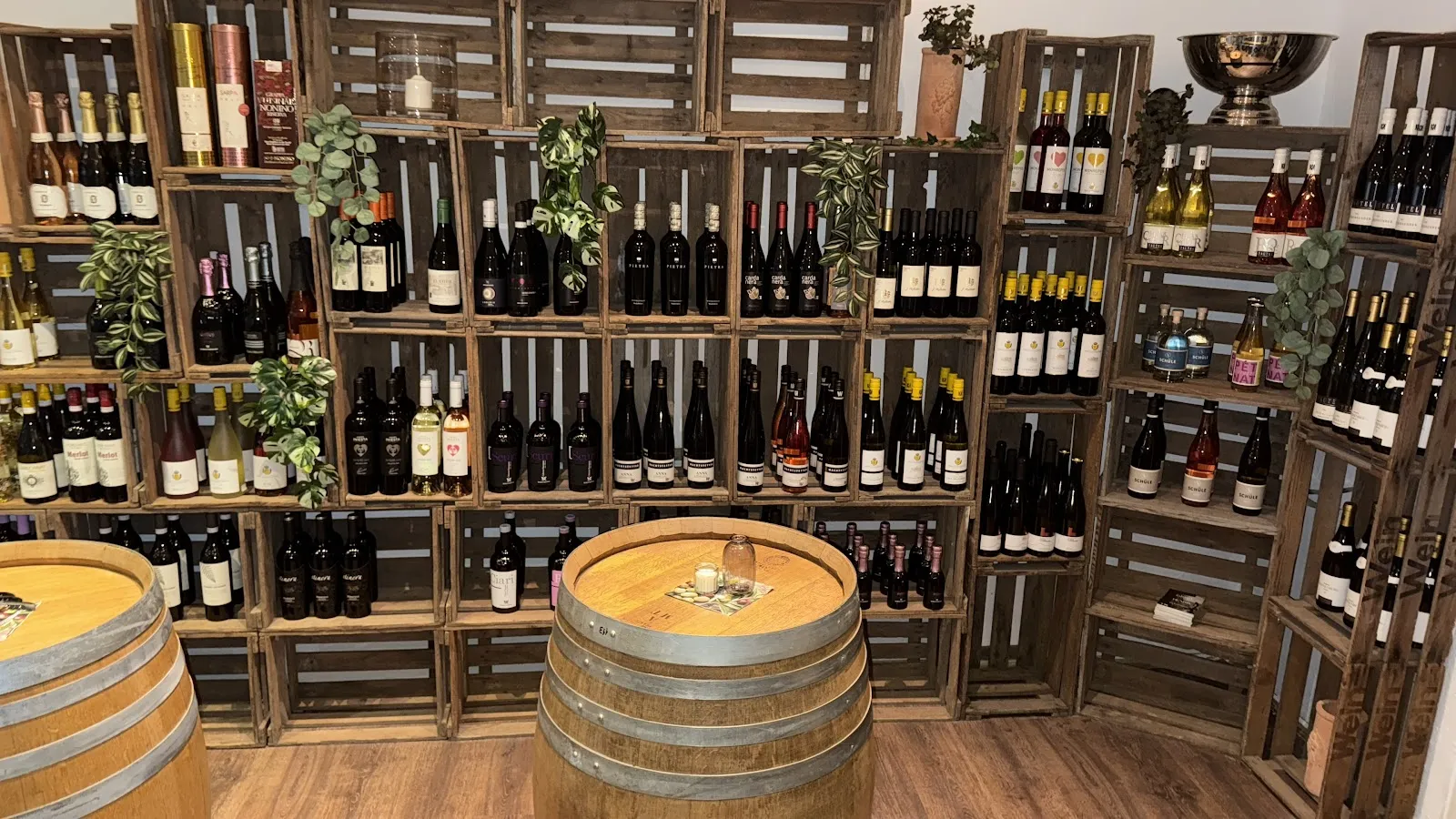Weinwand aus alten Holzkisten mit Flaschen und Grünpflanzen, davor Weinfässer als Stehtische.